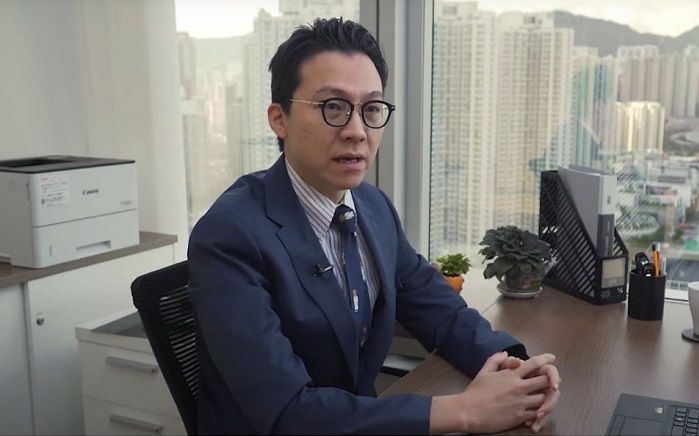 Dr. Lau seated at his desk with skyscrapers visible in the windows behind