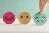 Red sad face, yellow neutral face, green smiley face held between a thumb and finger