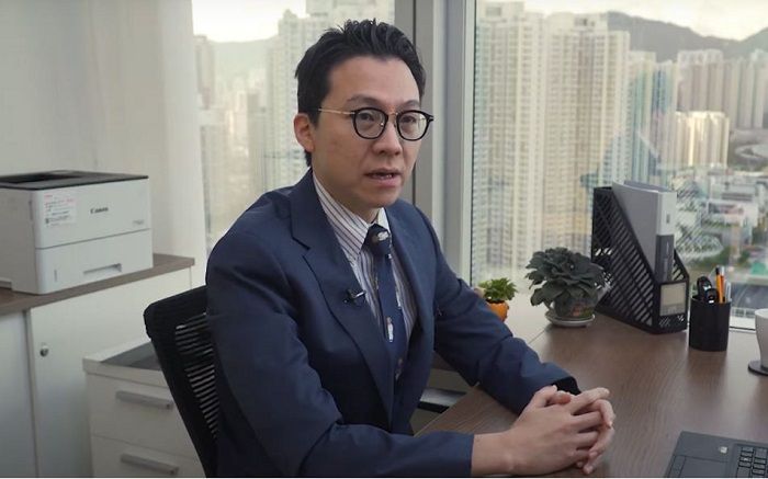 Dr. Lau seated at his desk with skyscrapers visible in the windows behind